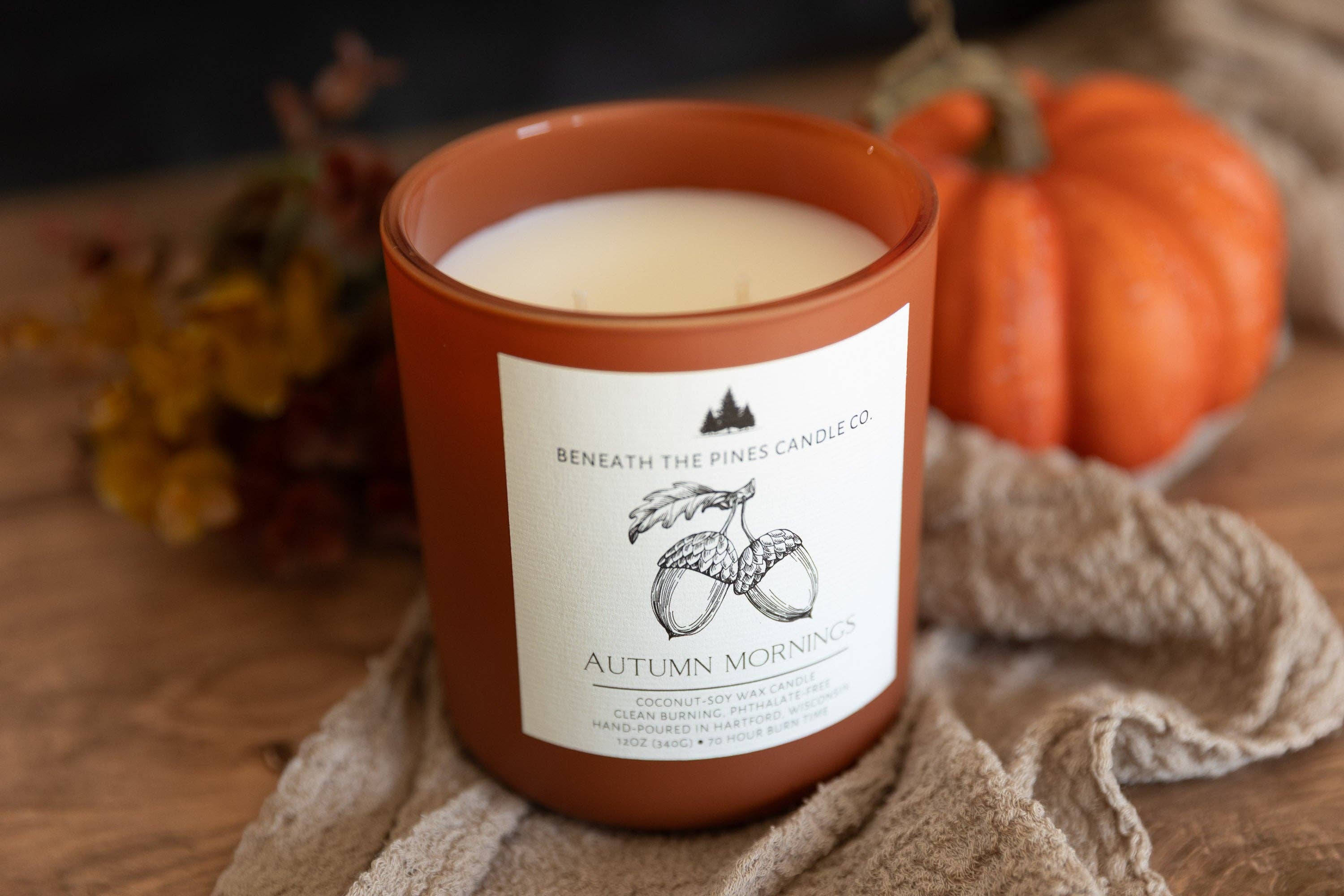 A terracotta-colored candle jar labeled 'Autumn Morning' by Beneath The Pines Candle Co, styled on a rustic wooden surface with a beige cloth, autumn leaves, and a small orange pumpkin in the background.