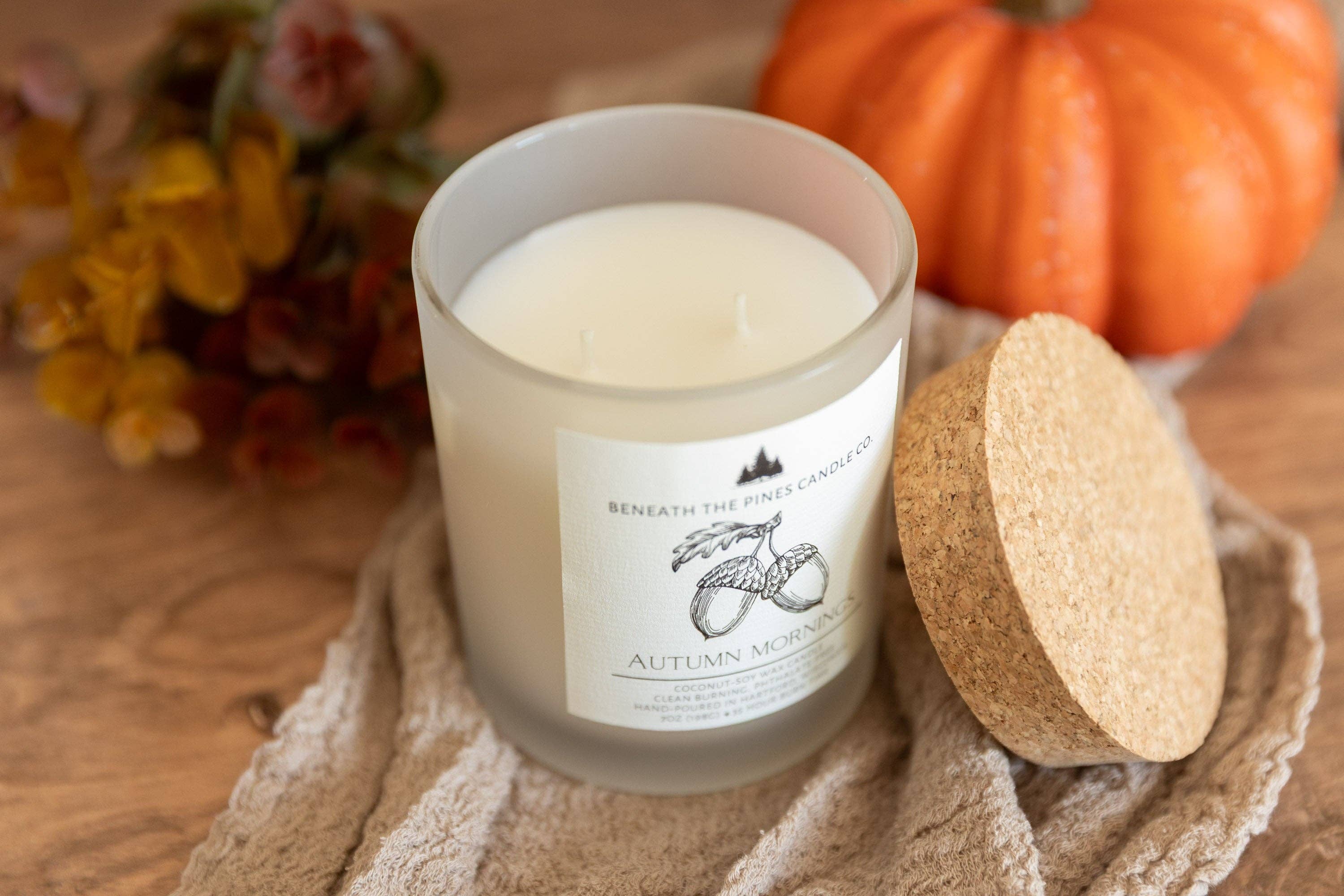 A frosted glass candle jar with a white wax fill and a white label reading 'Beneath the Pine Trees Candle' and 'Autumn Mood,' shown on a wooden surface with a cork lid beside it, autumn leaves in the background, and an orange pumpkin behind it.