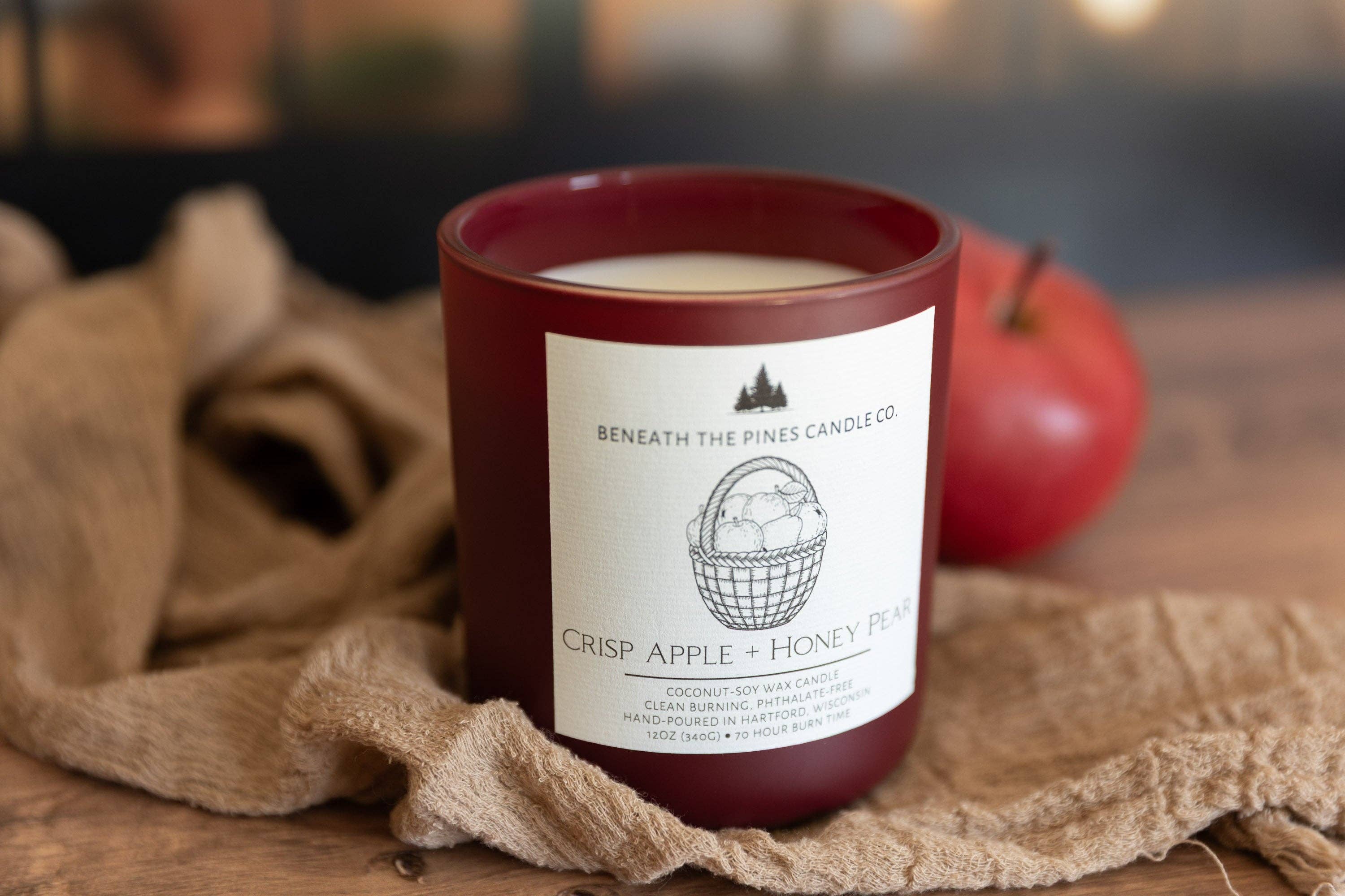 A dark red glass candle jar with a white label reading 'Beneath the Pines Candle Co. Crisp Apple + Honey Pine,' placed on a rustic wooden surface with burlap fabric and a red apple in the background.