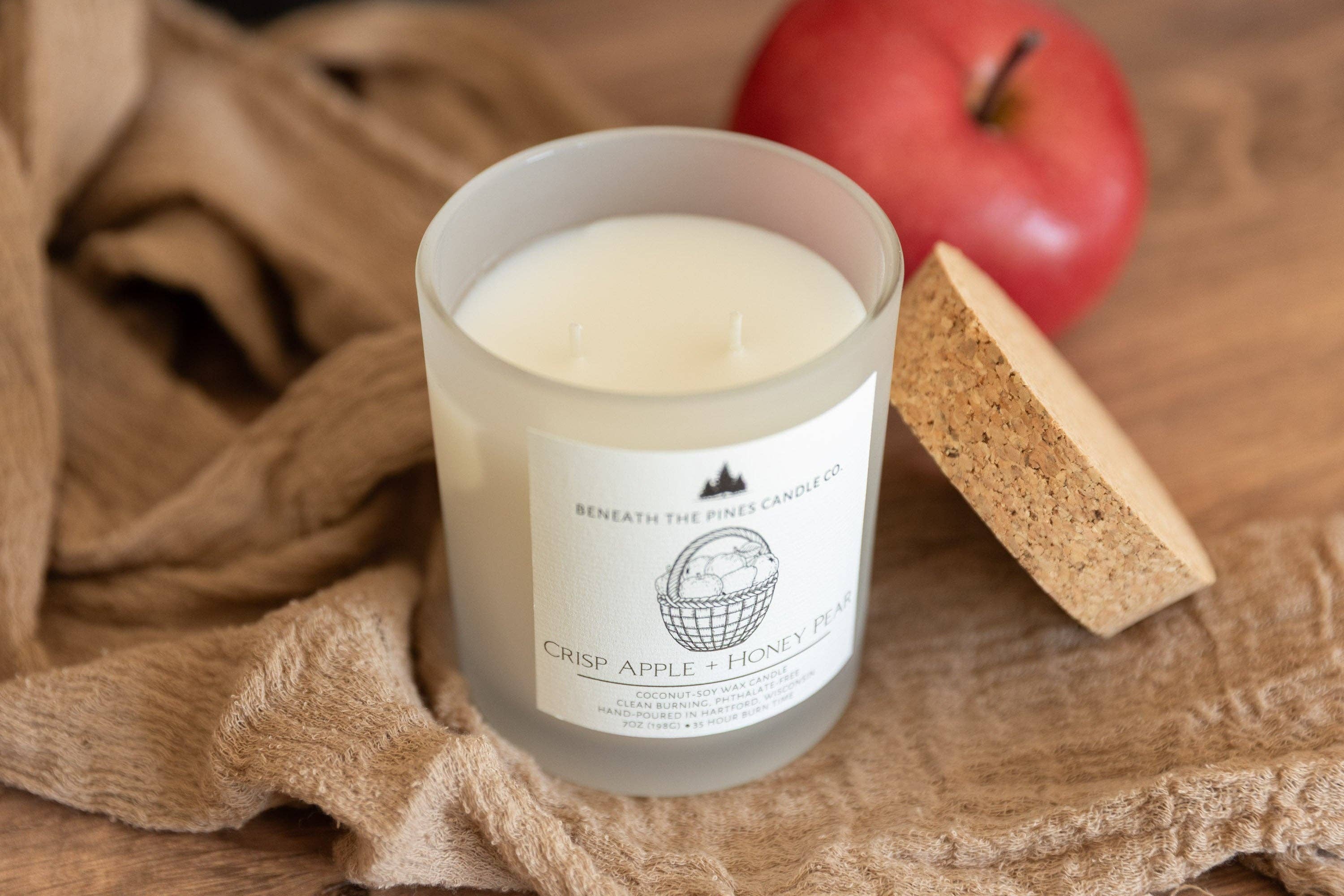 A frosted glass candle jar labeled 'Beneath the Pine Tree Co. Crisp Apple + Honey Balm' sits on a rustic wooden surface with a red apple and a cork wedge in the background.