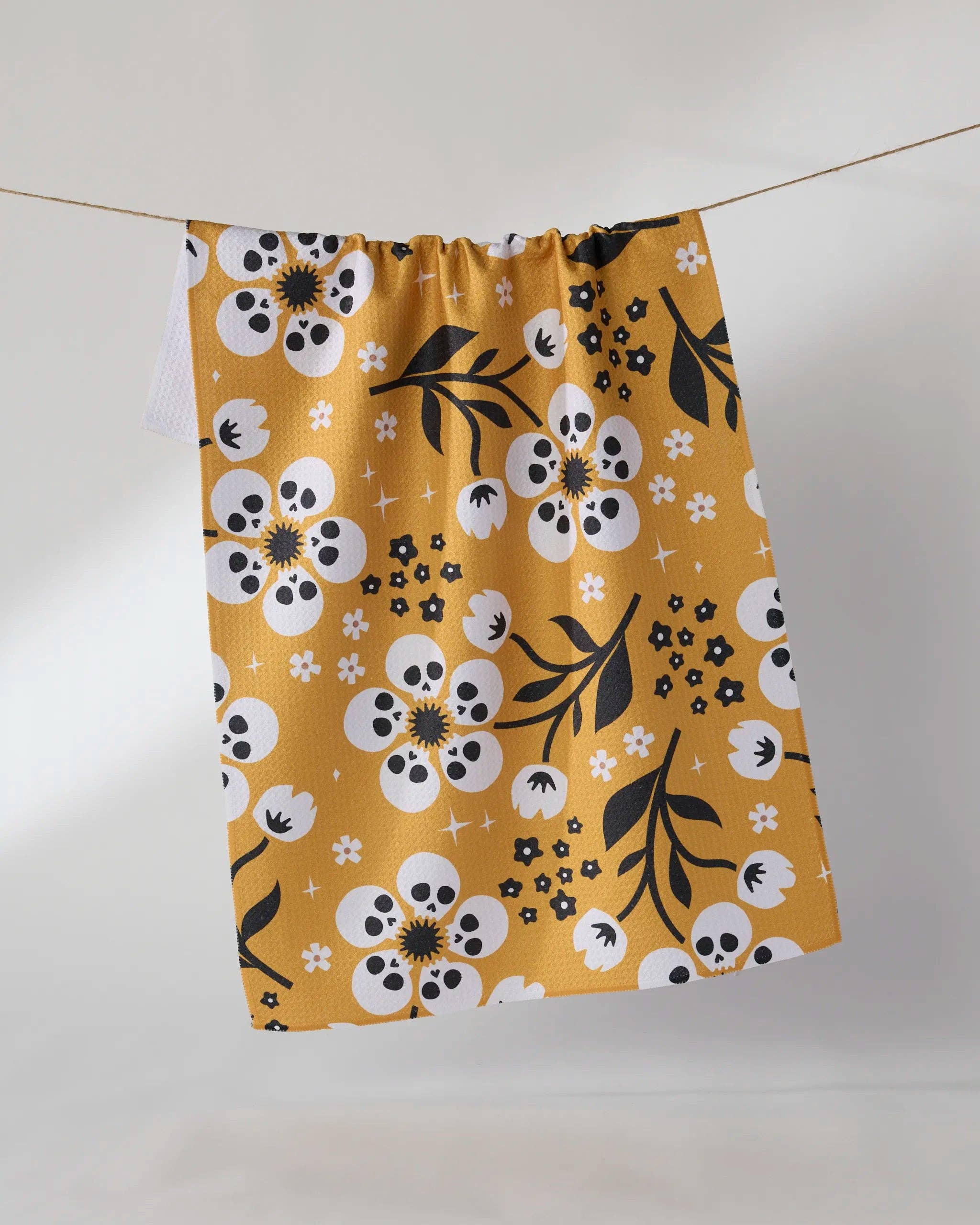A mustard yellow kitchen towel with a white and black floral and skull pattern, hanging from a string against a plain white background.