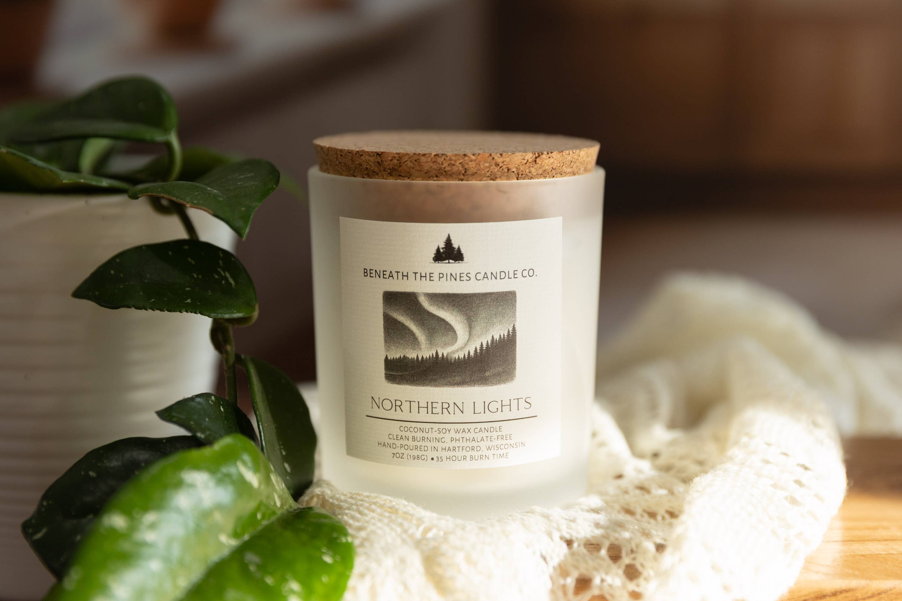 A frosted glass jar candle with a cork lid and a label reading 'Beneath the Pines Candle Co. Northern Lights,' styled on a wooden surface with a white lace cloth and green leaves in the foreground.