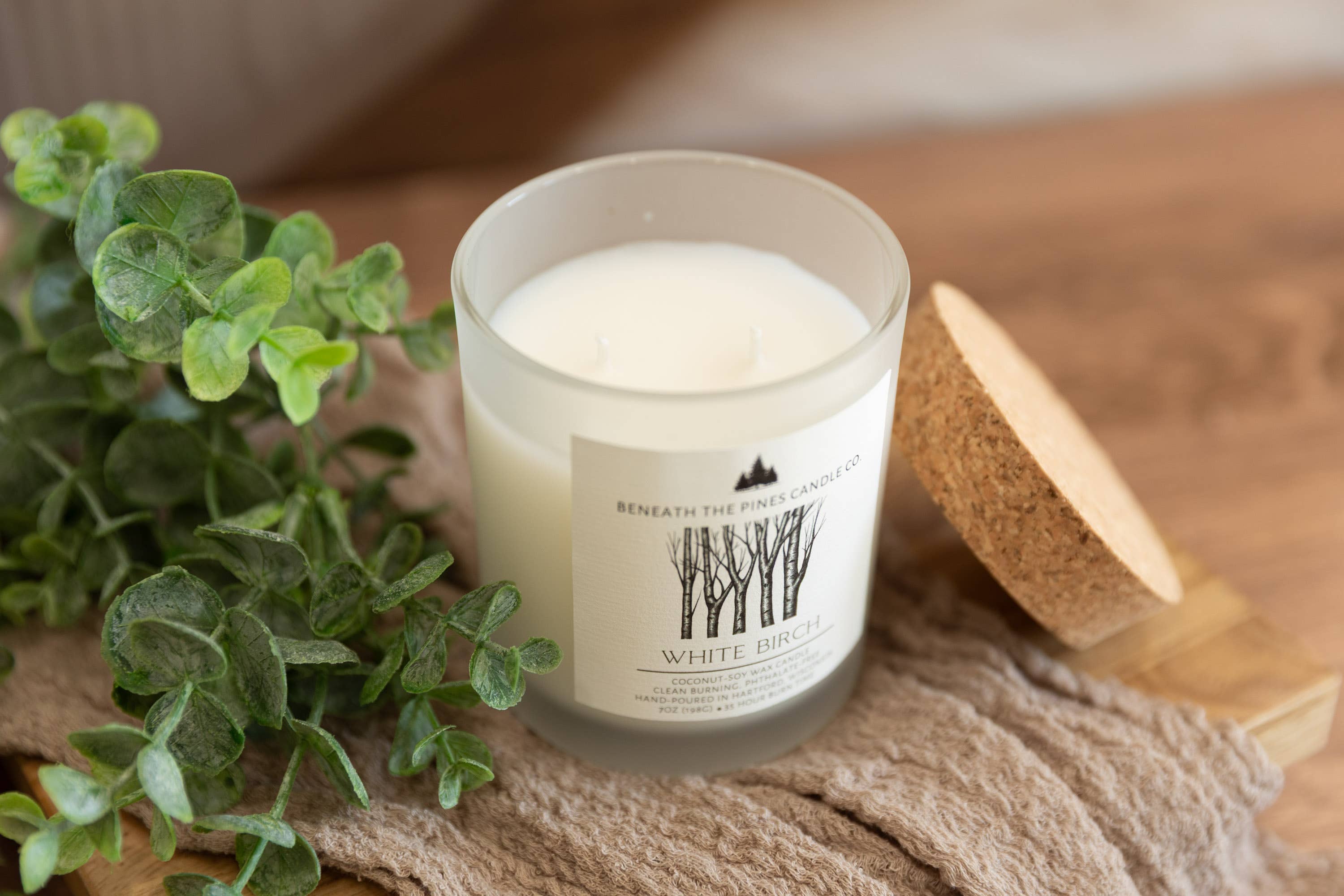 A white wax candle in a frosted glass jar with a white label reading 'Beneath the Pine Trees' and 'White Birch,' styled on a beige fabric surface with green eucalyptus leaves and a cork lid beside it.