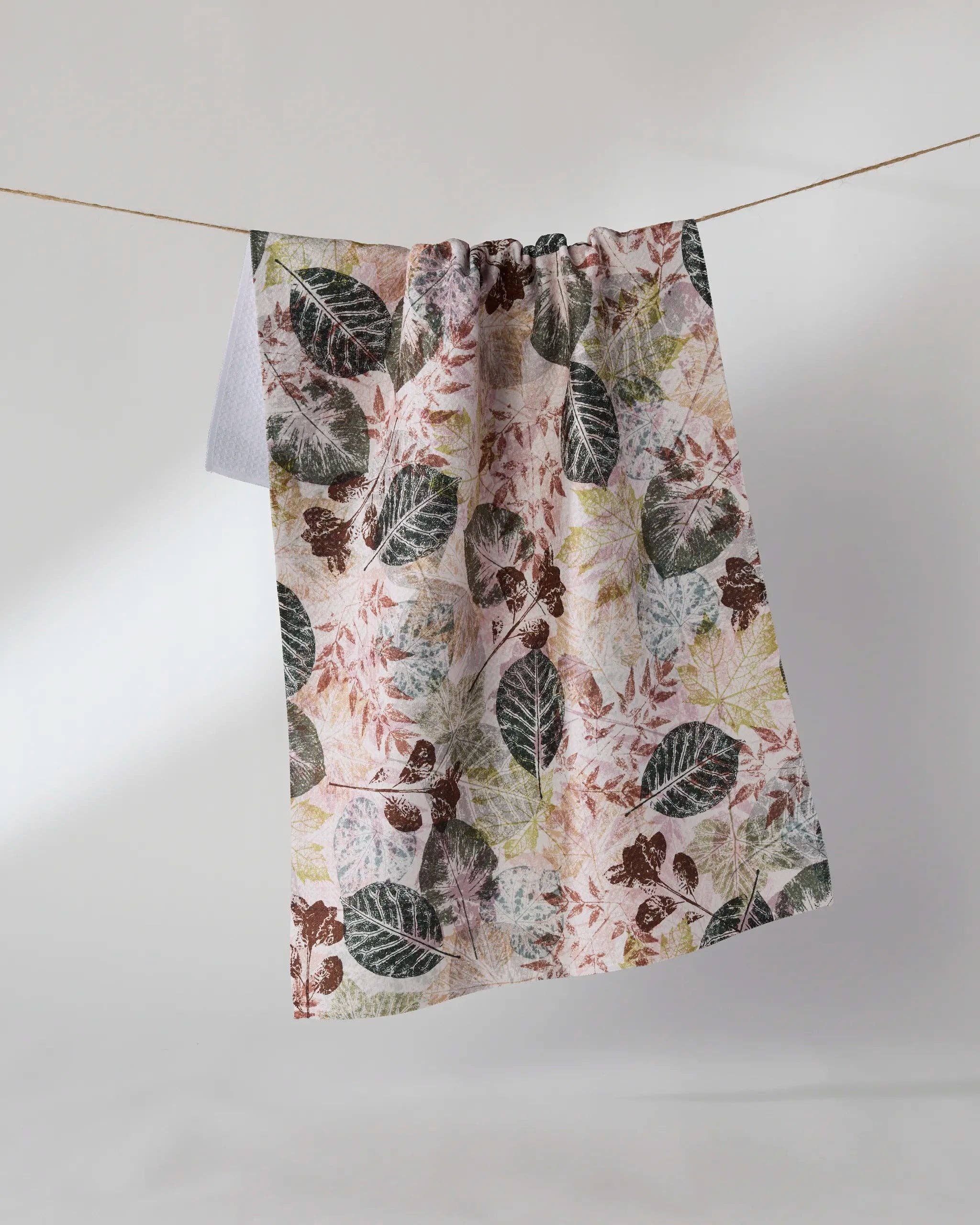 A lightweight fabric curtain panel with a botanical leaf pattern in muted green, brown, and dusty pink on a cream background, hanging from a thin wire against a plain light gray studio backdrop.