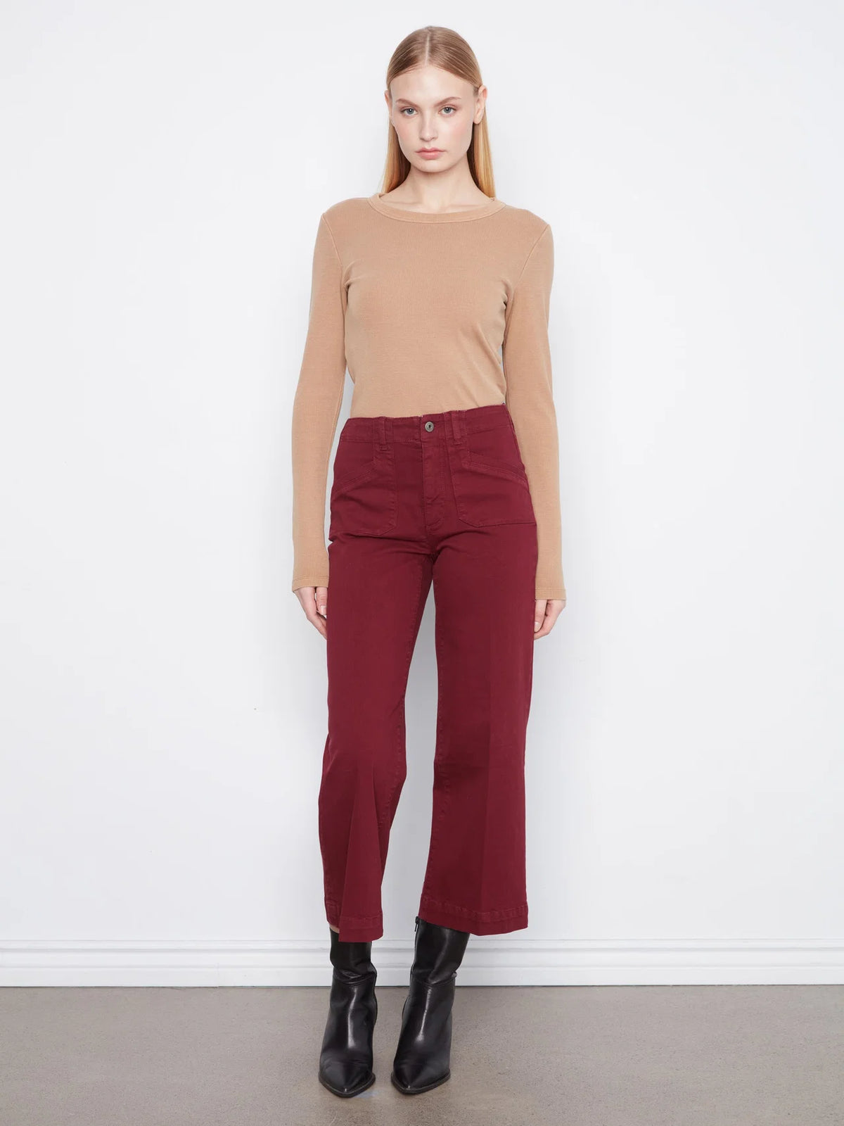 A woman stands against a plain white wall wearing a fitted tan long-sleeve top, dark red cropped pants, and black ankle boots.
