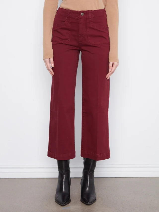 A model wearing high-waisted, wide-leg cropped pants in a deep burgundy red, paired with a beige long-sleeve top and black leather ankle boots, standing against a white wall.