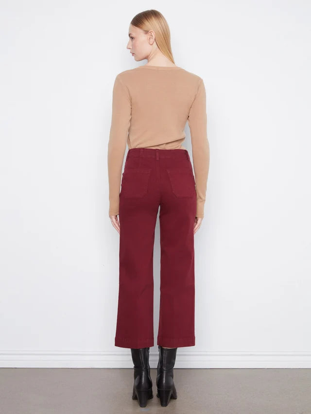 A woman stands with her back to the camera wearing a beige long-sleeve top, dark red wide-leg cropped pants, and black ankle boots against a plain white wall.