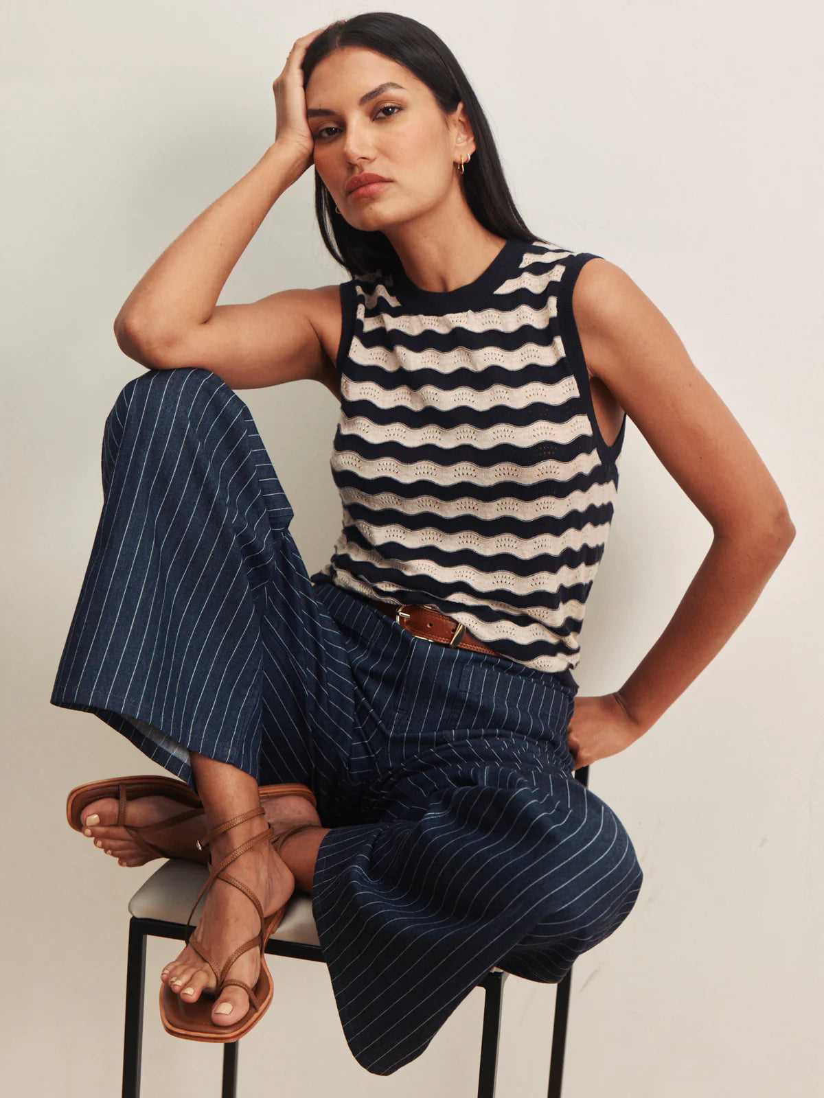 Woman wearing a striped sleeveless top and pinstripe pants sitting on a chair.