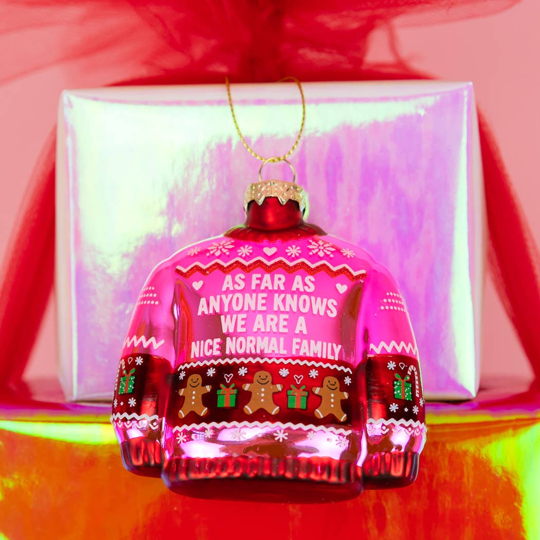 Pink glass ornament by Chez Gagné shaped like an ugly sweater, reads As Far As Anyone Knows We Are A Nice Normal Family