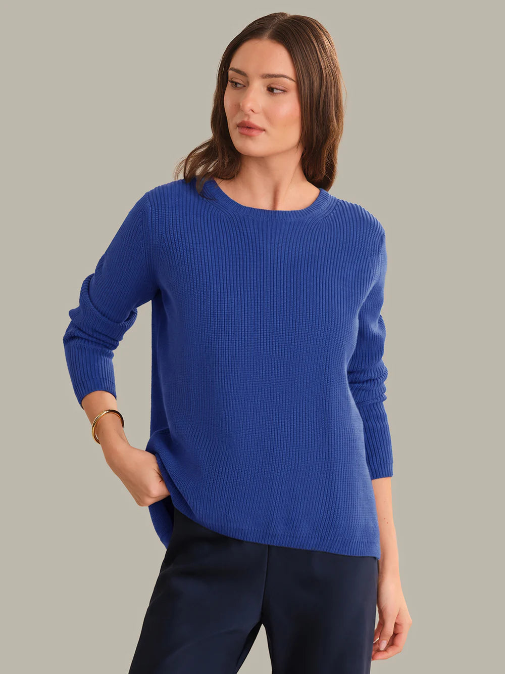 A woman wearing a royal blue ribbed knit sweater and dark navy trousers, standing against a solid light gray background.