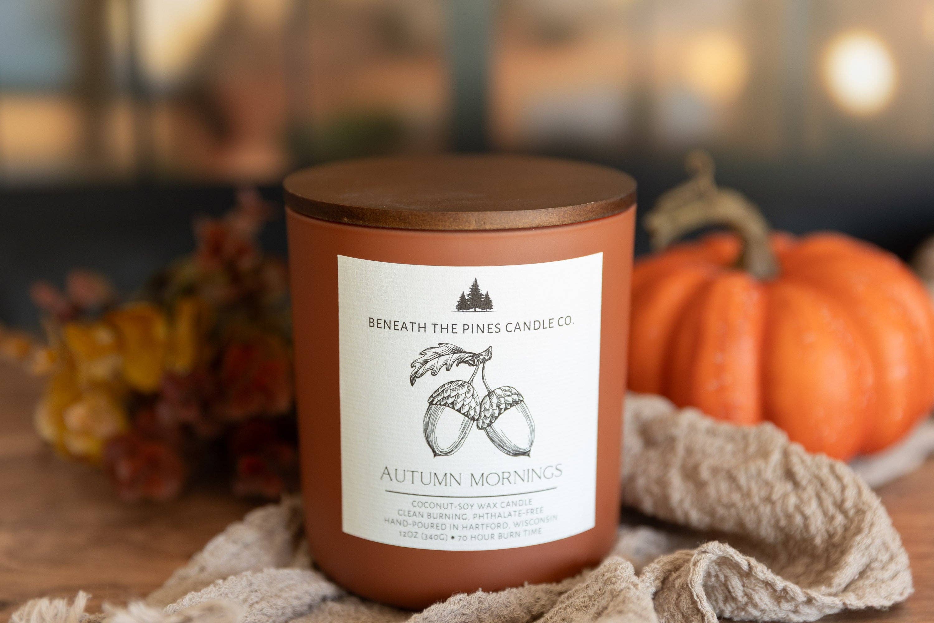 A candle jar labeled 'Beneath the Pines Candle Co. Autumn Mornings' with a warm terracotta body, a dark wood lid, and a white front label, styled on a table with autumn leaves, a pumpkin, and a textured cloth.