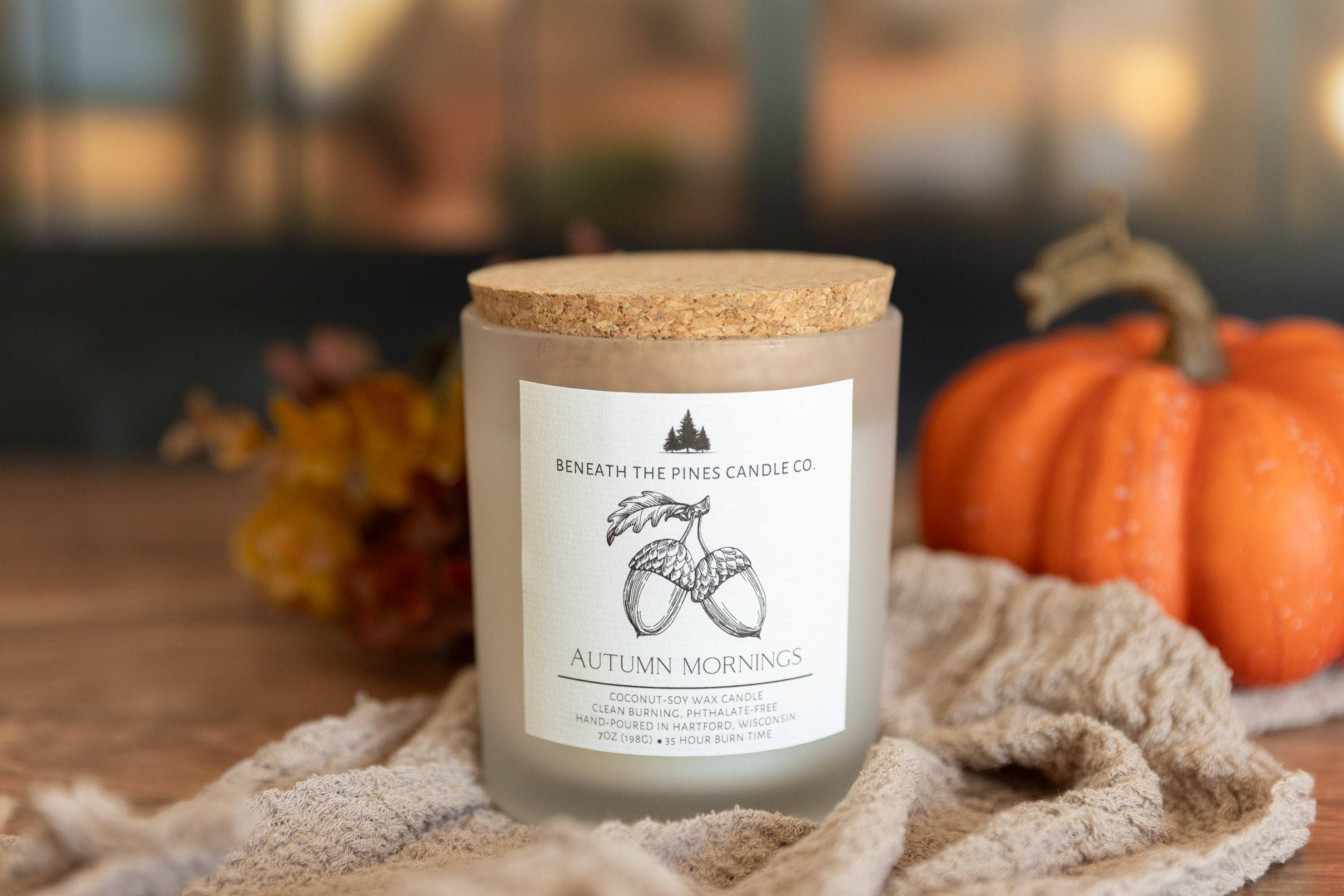 A frosted glass jar candle with a cork lid and a white label reading 'Beneath the Pines Candle Co. Autumn Mornings,' styled on a textured beige towel with pumpkins and autumn leaves in a warm, softly blurred background.