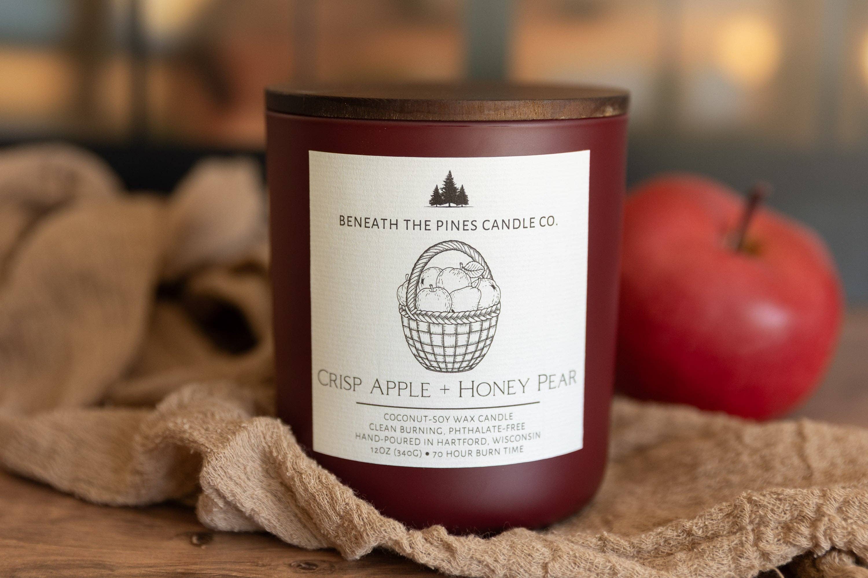 A dark red soy wax candle by Beneath The Pines Candle Co. labeled 'Crisp Apple + Honey Pear,' shown on a rustic wooden surface with burlap and a red apple in the background.