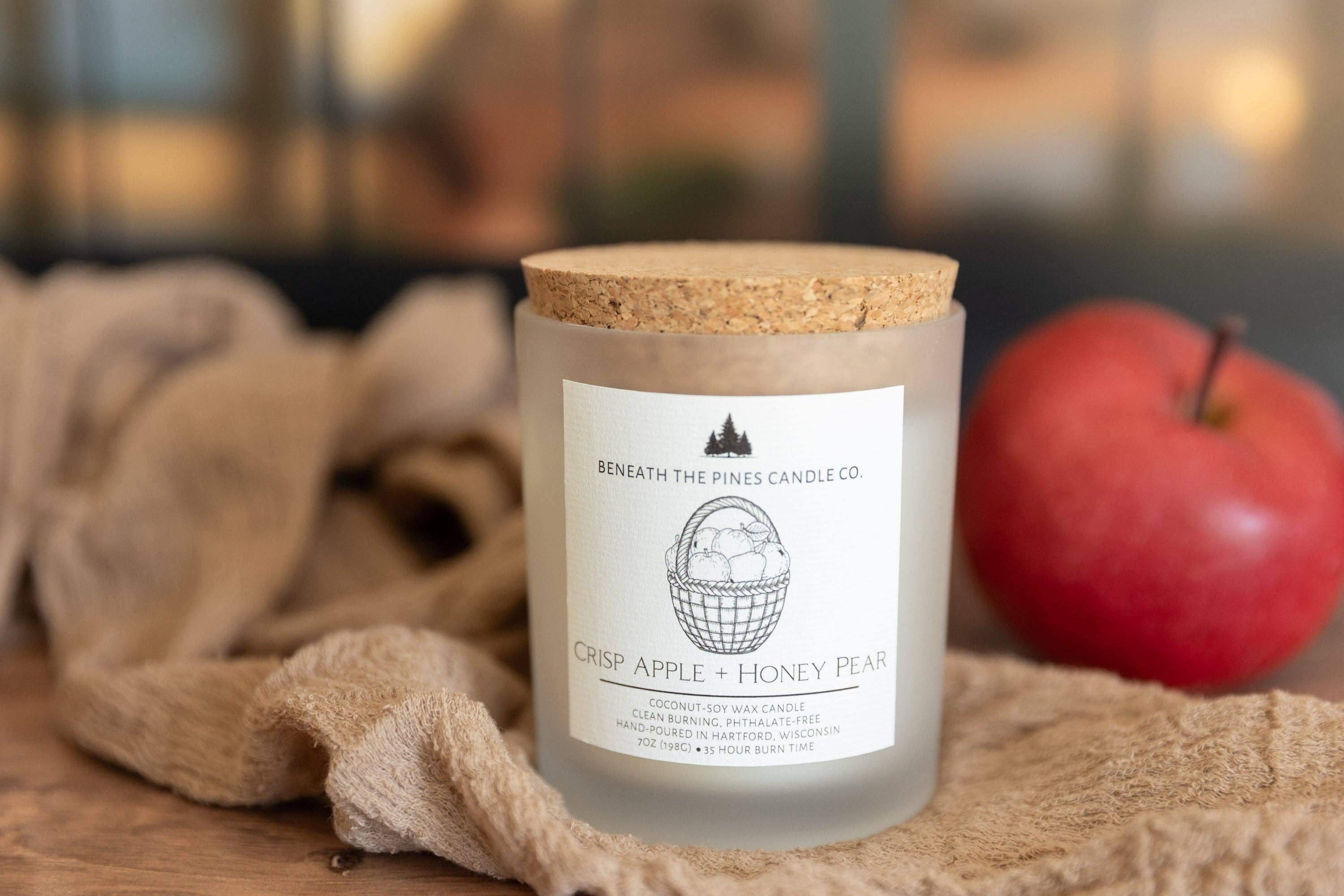 A frosted glass jar candle with a cork lid sits on a wooden surface next to a beige cloth and a red apple. The front label reads 'BENEATH THE PINES CANDLE CO' and 'CRISP APPLE + HONEY PEAR'.