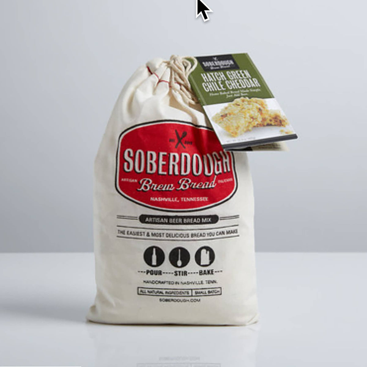 A beige fabric drawstring bread bag labeled Soberdough Brien Bread, with a green and white hanging tag attached near the top, photographed against a clean light gray studio background.