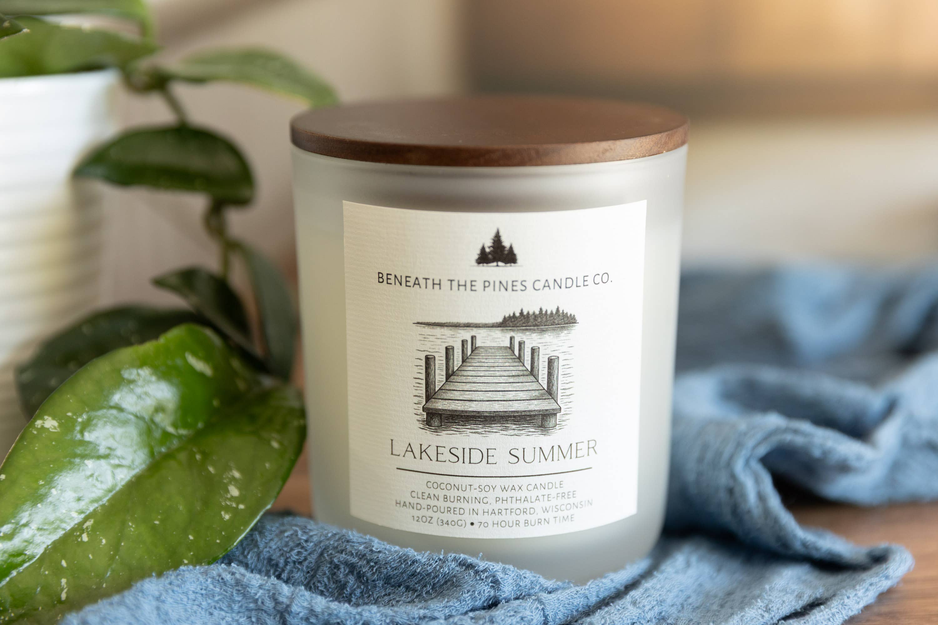 A frosted glass candle jar with a wooden lid and a cream label reading 'Beneath The Pines Candle Co. Lakeside Summer,' styled on a blue cloth with green leaves in the foreground.