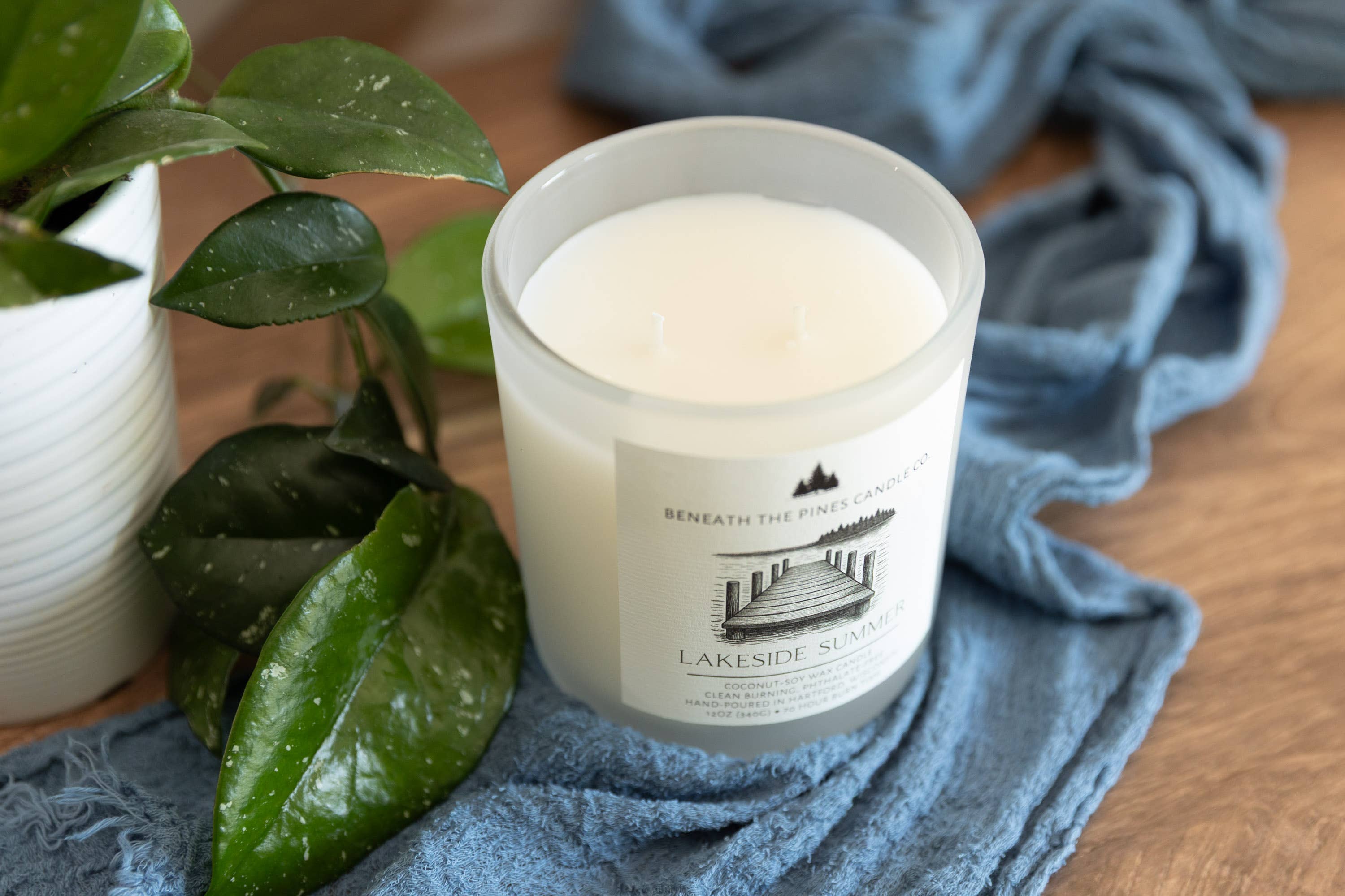 A frosted glass jar candle with white wax sits on a blue textured cloth on a wooden surface, styled with green leaves and a white planter.