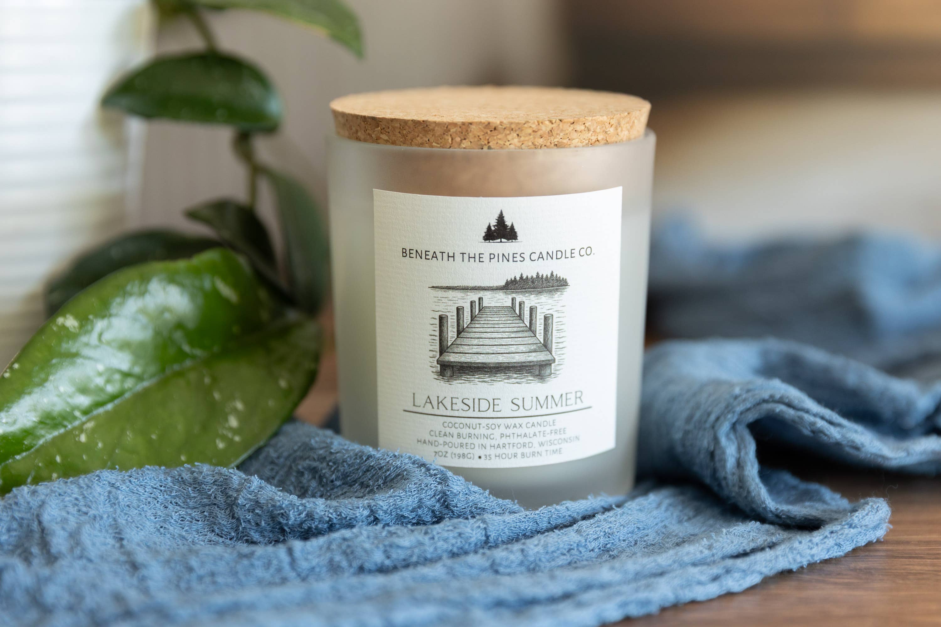 A glass jar candle with a cork lid and a white label reading 'Beneath the Pines Candle Co. Lakeside Summer,' styled on a blue textured cloth with green leaves in the background.