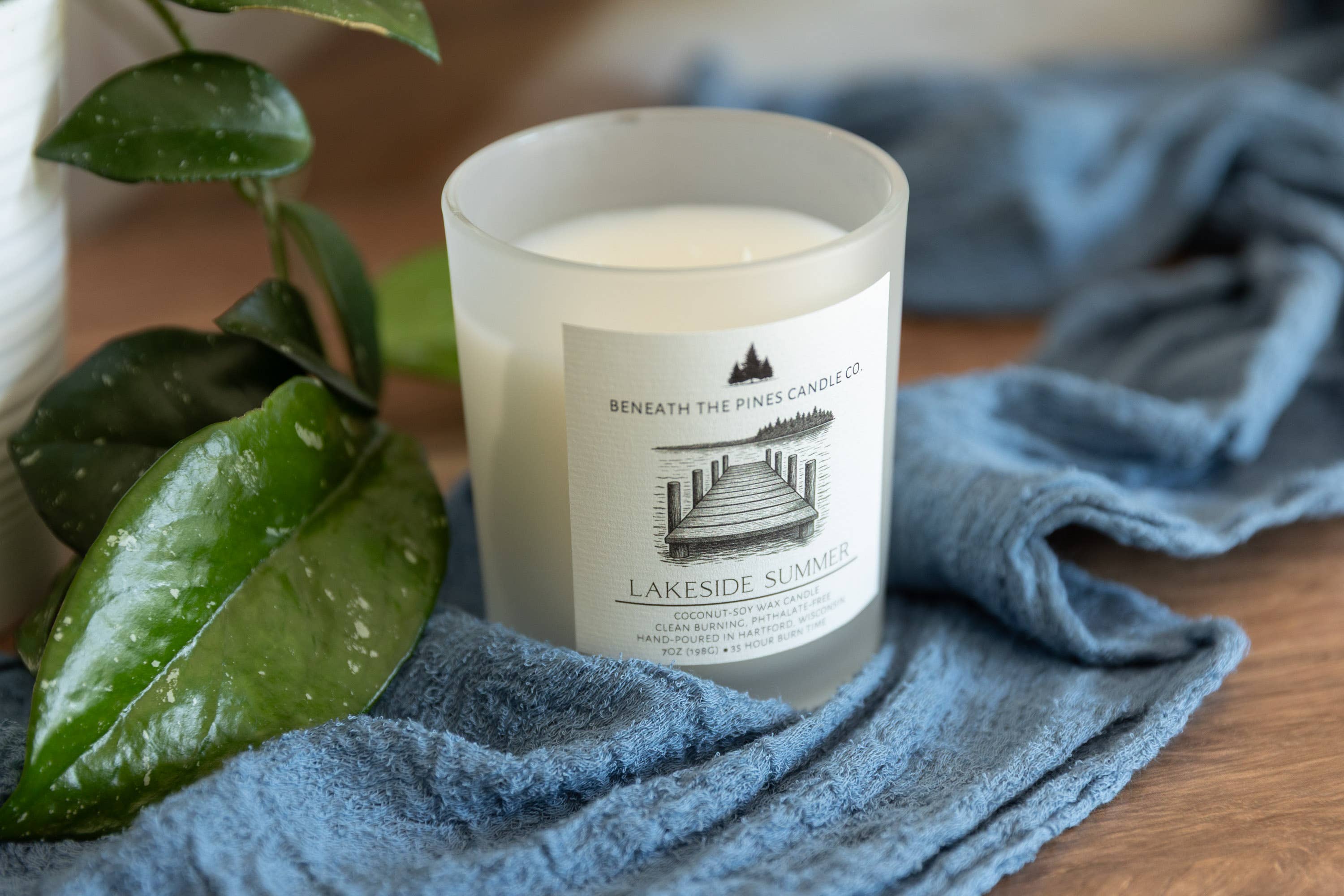 A frosted glass candle jar labeled 'Beneath the Pine Candle Co. Lakeside Summer' sits on a blue textured cloth over a wooden surface, with green leaves entering from the left.