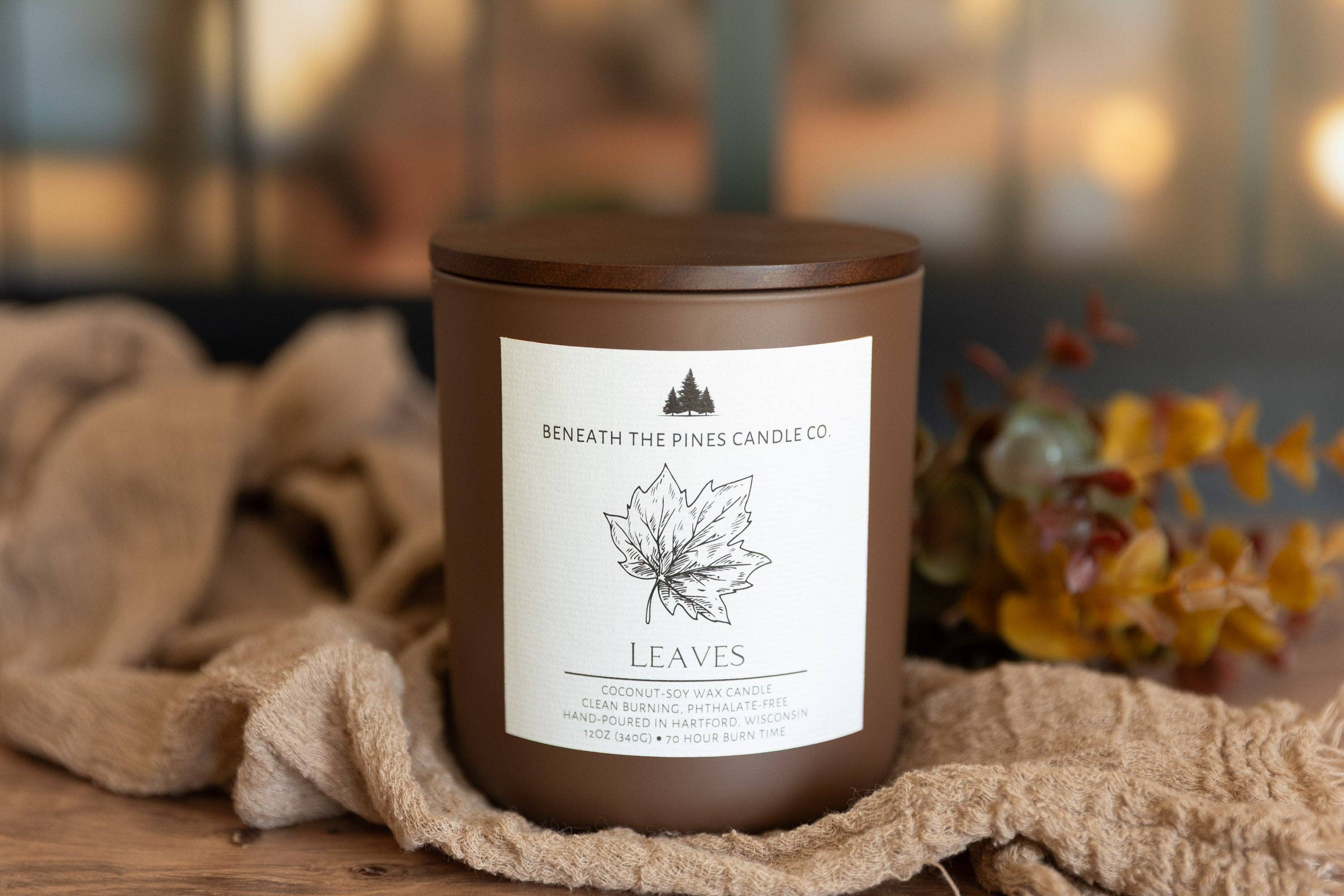 A brown glass jar candle with a matching lid and a white label reading 'Beneath the Pines Candle Co.' and 'LEAVES,' styled on a wooden surface with beige fabric and blurred autumn flowers in the background.