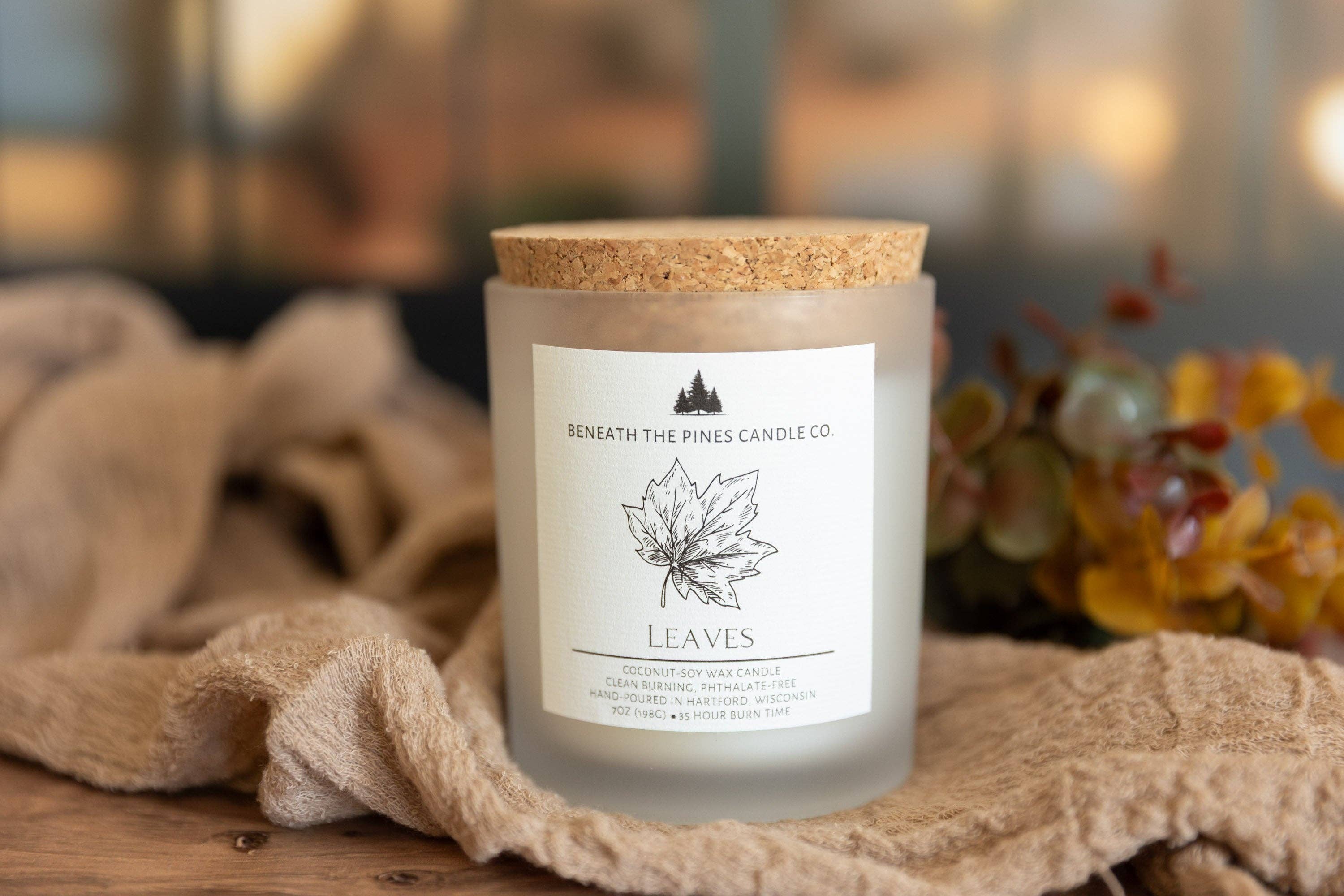 A frosted glass candle jar with a cork lid and a white label reading 'Beneath The Pines Candle Co.' and 'LEAVES,' styled on a wooden surface with a beige cloth and blurred autumn foliage in the background.