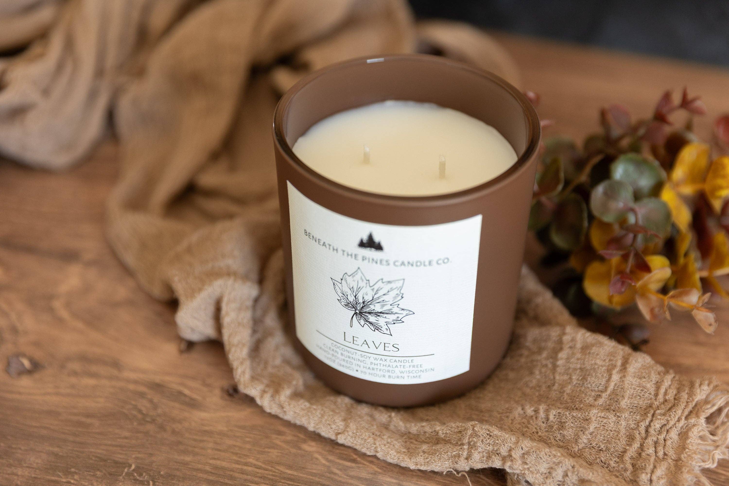 A brown glass jar candle with a white label reading 'Majestic The Pine Candle Co' and 'LEAVES', placed on a rustic wooden surface with a beige fabric and dried flowers in the background.