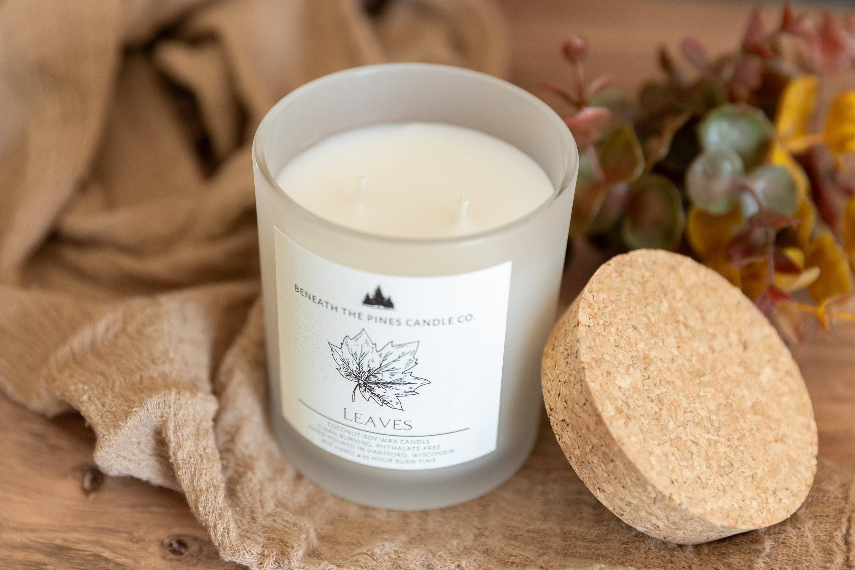 A frosted glass candle jar with white wax and a white label reading 'Meadath the Pine Candle Co' and 'LEAVES', shown on a wooden surface with a cork lid beside it and soft decorative fabric and succulents in the background.