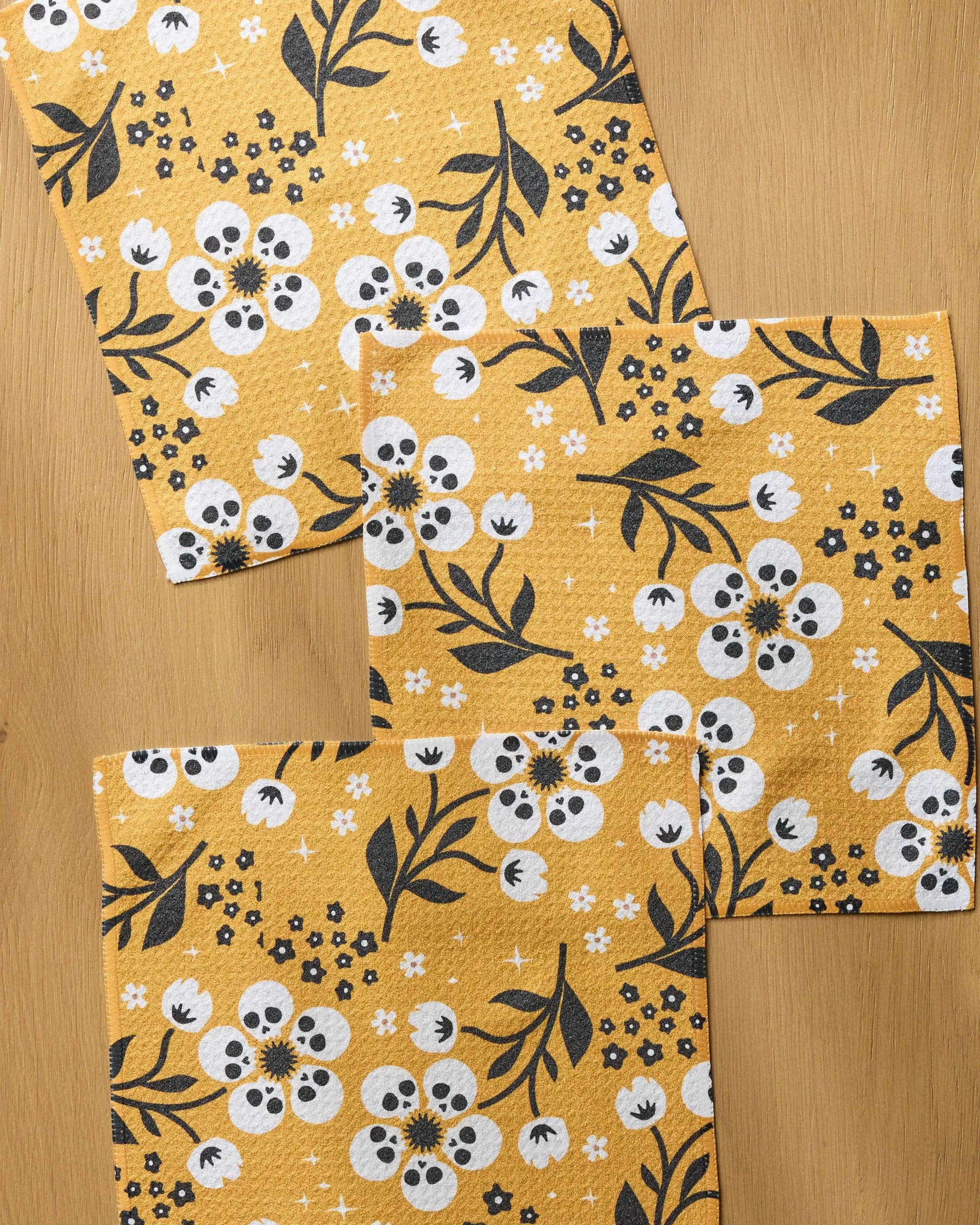 Three square kitchen napkins with a yellow, white, and black floral pattern arranged on a light wood surface.