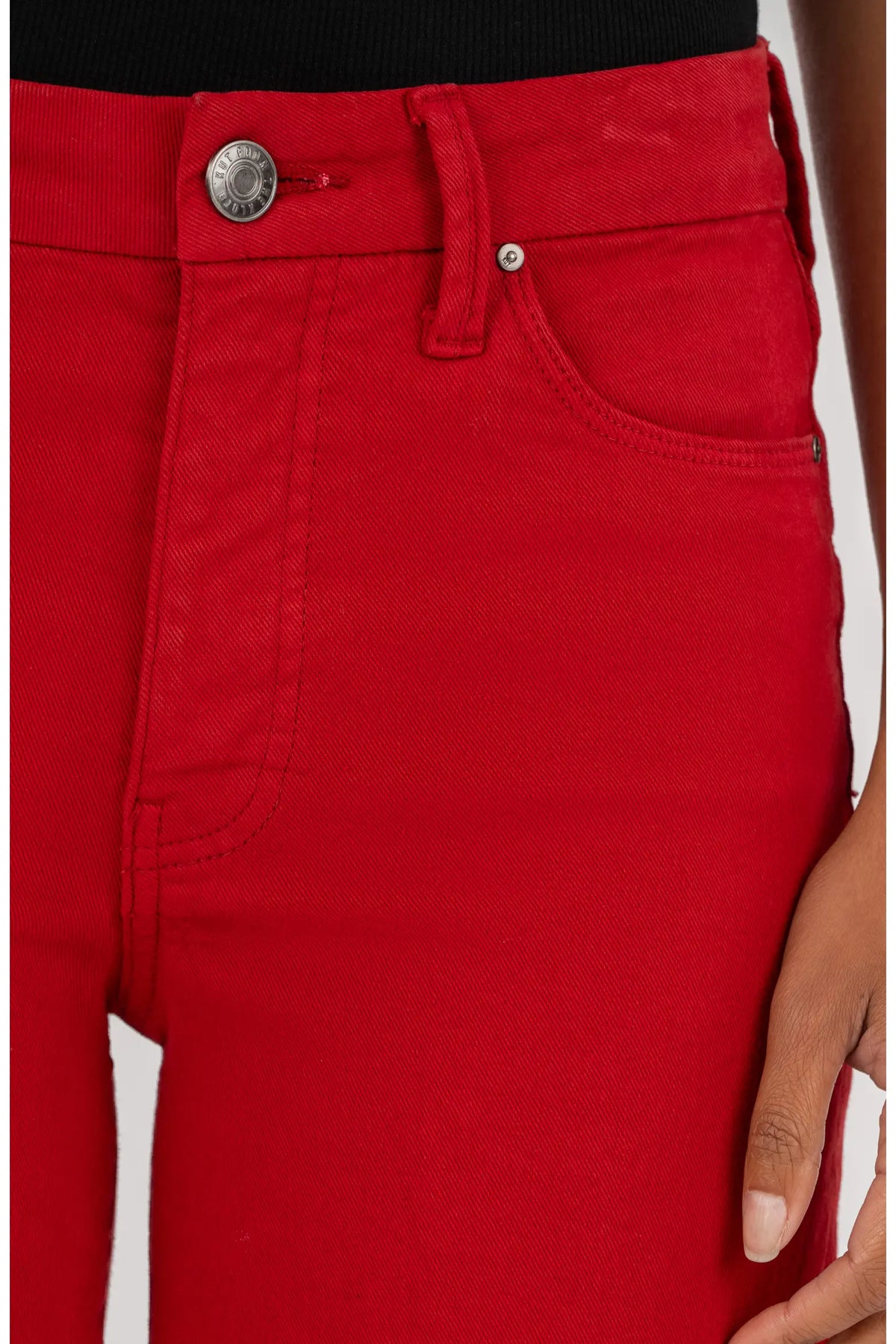 Close-up of bright red denim jeans worn by a model, showing the front waistband with a silver-tone button, belt loop, pocket, and part of the hand holding the hem.