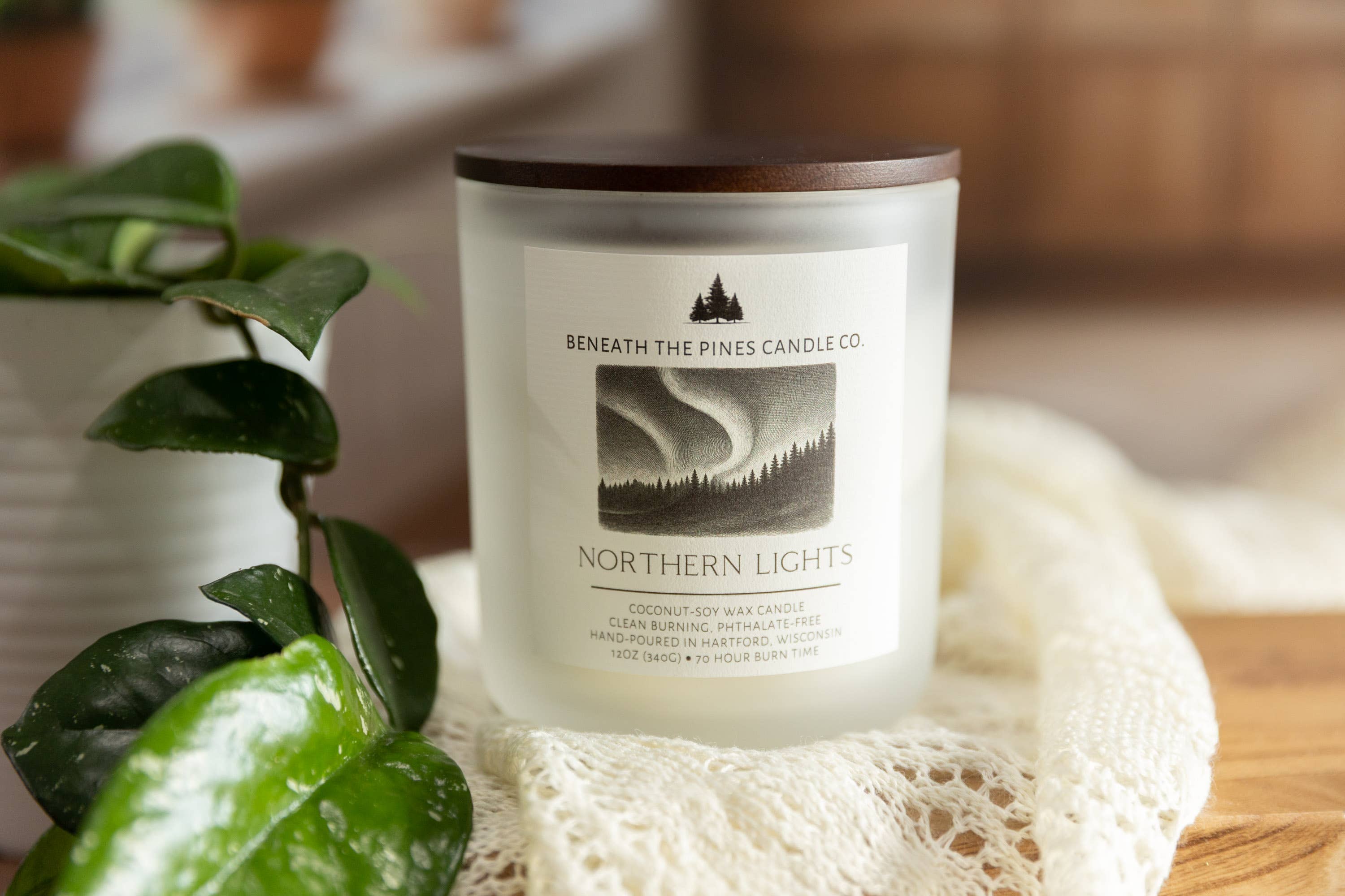 A jar candle labeled 'Northern Lights' by Beneath The Pines Candle Co., shown with a white wax body, dark wood lid, and a cream lace cloth on a wooden surface with green leaves in the foreground.