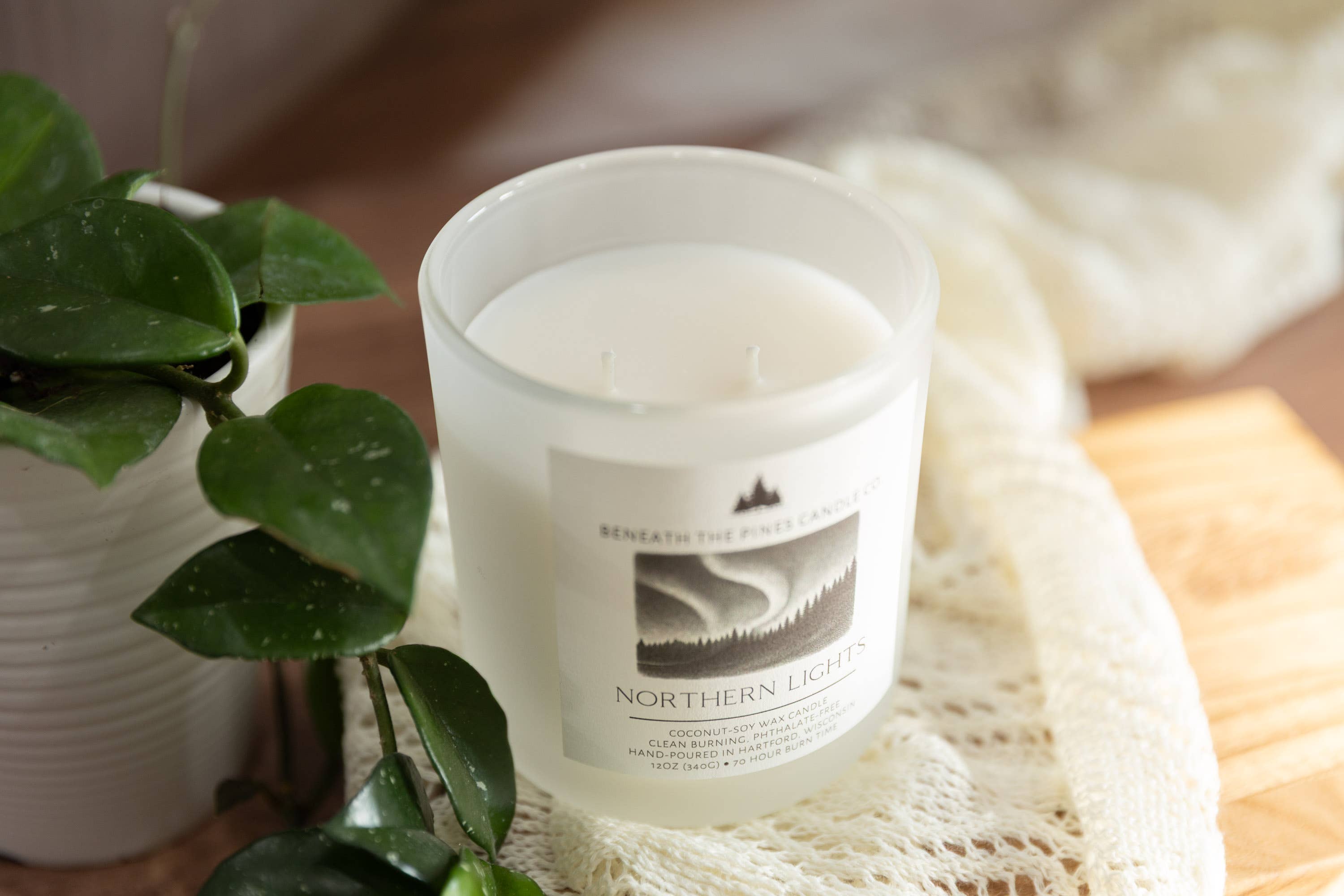 A frosted glass candle jar labeled 'Northern Lights' by Beneath the North Light Candle, styled on a cream lace cloth next to a potted green plant.
