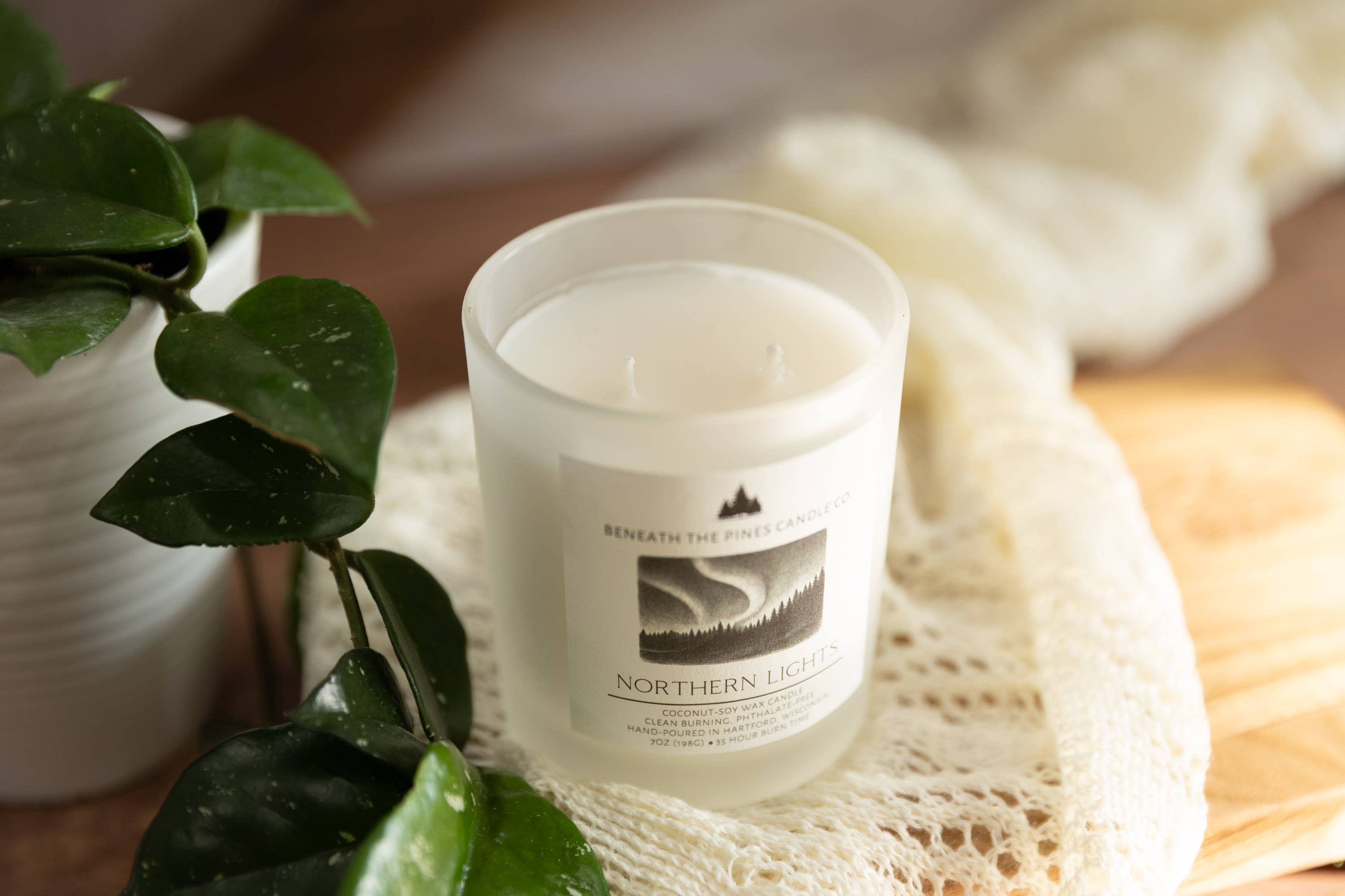 A frosted glass two-wick candle labeled 'Northern Lights' by 'Beneath the Northern Lights' sits on a cream lace cloth next to green plant leaves.