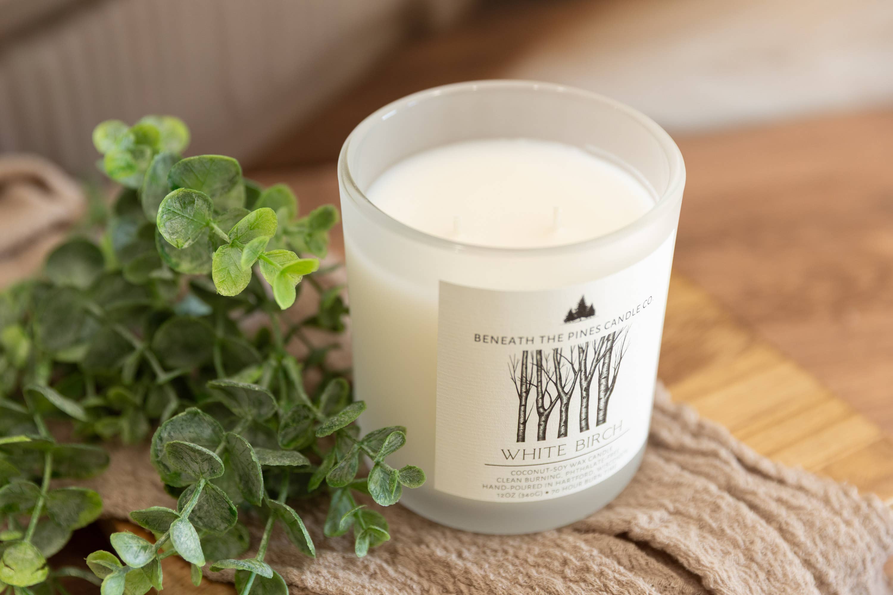 A glass jar candle with white wax sits on a beige textured blanket beside green eucalyptus leaves on a wooden surface. The label reads 'Beneath the Pine Candles' and 'White Birch'.