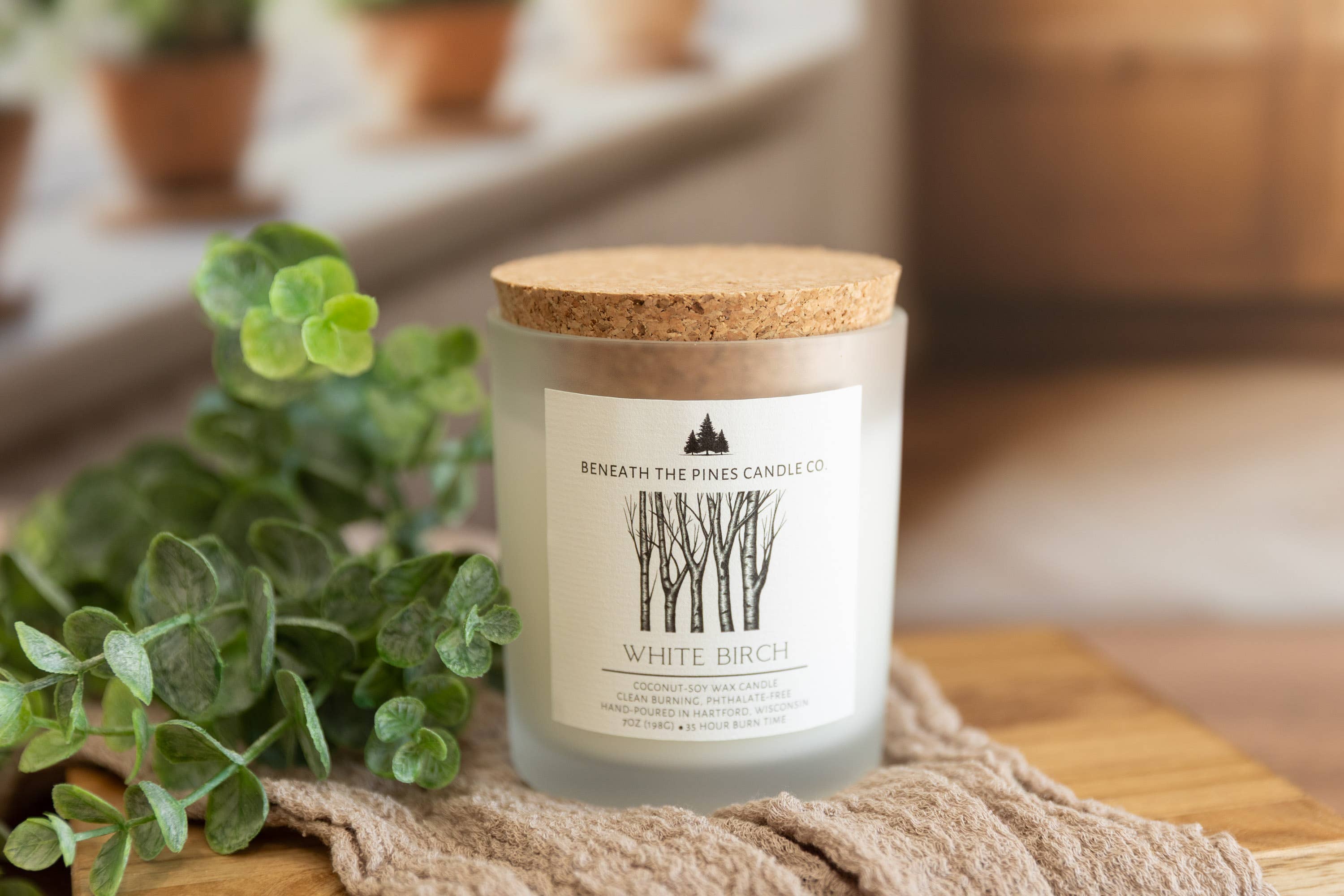 A glass jar candle with a cork lid and a white label reading 'Beneath The Pines Candle Co' and 'White Birch', styled on a wooden surface with a beige cloth and green leaves in a softly blurred indoor setting.