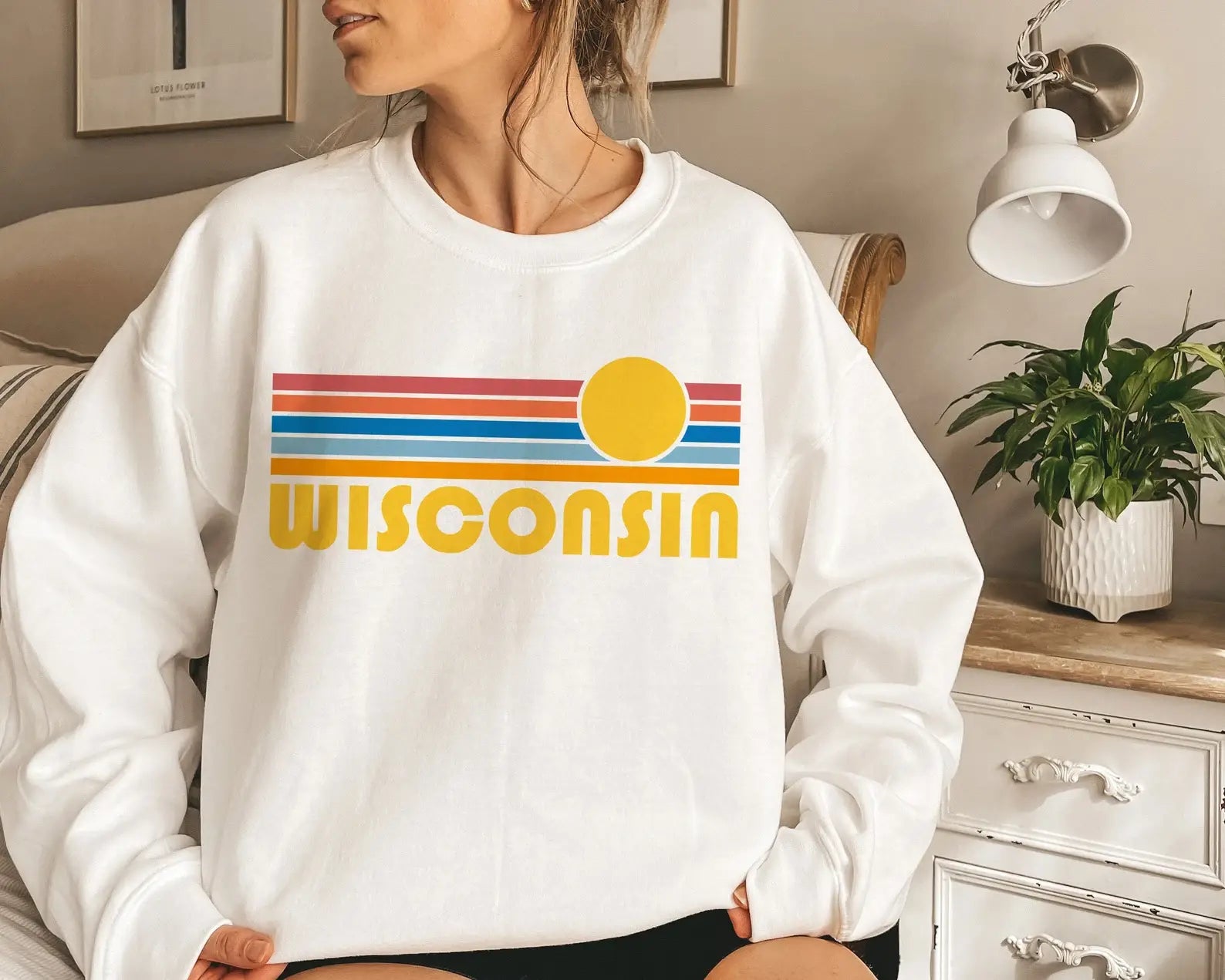 A woman wearing a white crewneck sweatshirt with a retro-style 'Wisconsin' graphic featuring horizontal red, blue, and orange stripes, a yellow sun, and yellow text, shown in a cozy bedroom setting.
