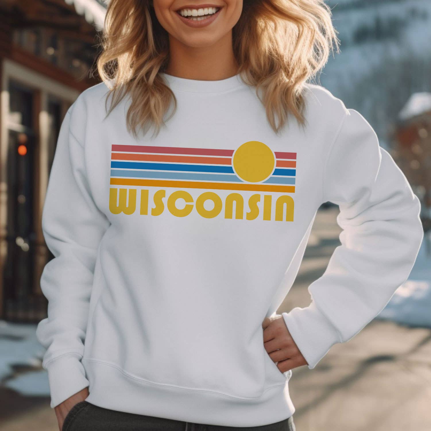 A woman wearing a white crewneck sweatshirt with a retro-style 'Wisconsin' graphic featuring yellow text and orange, blue, and red stripes with a yellow sun, set against a blurred outdoor background.